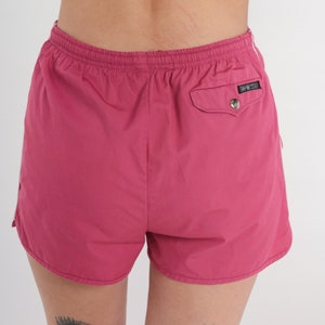 May include: Magenta athletic shorts with a white stripe down the side. The shorts feature an elasticated waistband and a small pocket on the back with a silver snap closure. Made from a lightweight material, suitable for sports.