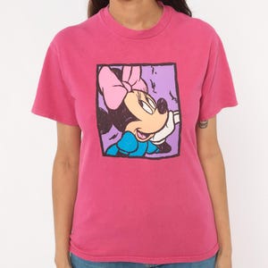 May include: A short-sleeved crew neck t-shirt in a vibrant pink colour. The centre of the shirt features a square graphic print of a cartoon mouse character with a pink bow, wearing a blue top, set against a light purple background with black outlines.