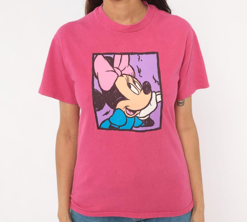 May include: A short-sleeved crew neck t-shirt in a vibrant pink colour. The centre of the shirt features a square graphic print of a cartoon mouse character with a pink bow, wearing a blue top, set against a light purple background with black outlines.