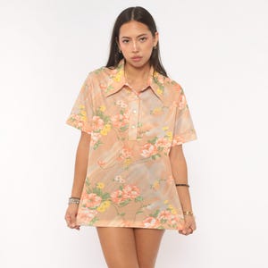 May include: A short-sleeved collared shirt in a light peach hue featuring a repeating floral pattern of pink and yellow blossoms with green leaves. The shirt has a button-up placket with three visible white buttons and a straight hemline.