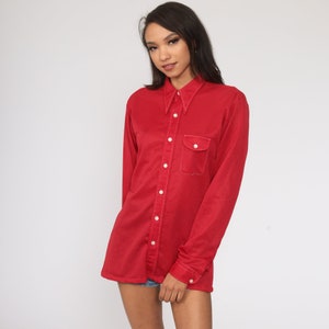 May include: A red corduroy shirt with a pointed collar, long sleeves, and a single chest pocket. The shirt has white buttons down the front and on the cuffs. The fabric has a ribbed texture.