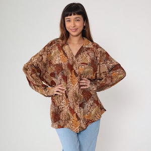 May include: A button-down blouse with a V-neck and long sleeves. The blouse features a brown, tan, and rust-colored pattern. The model is wearing light blue jeans. The blouse is made of a lightweight fabric.