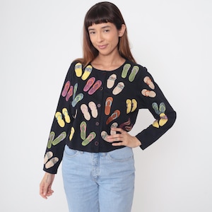May include: Black button-down cardigan sweater with colorful embroidered flip-flop designs. The long-sleeved sweater features a round neckline and a button-up front. The flip-flops are in various colors, including yellow, pink, green, and orange.