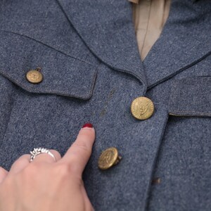 K&ouml;nnte beinhalten: Nahaufnahme einer blauen Wolljacke mit goldenen Kn&ouml;pfen. Die Jacke hat eine taillierte Passform, einen Reverskragen und zwei Vordertaschen. Die Kn&ouml;pfe haben ein detailliertes Emblem. Die Jacke scheint Vintage zu sein.