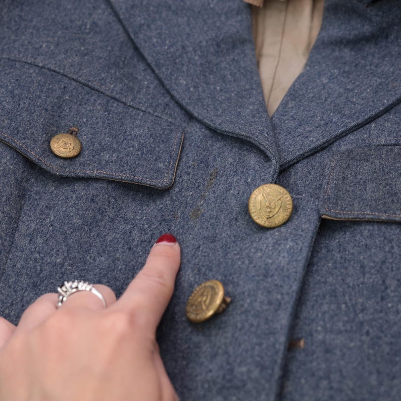 K&ouml;nnte beinhalten: Nahaufnahme einer blauen Wolljacke mit goldenen Kn&ouml;pfen. Die Jacke hat eine taillierte Passform, einen Reverskragen und zwei Vordertaschen. Die Kn&ouml;pfe haben ein detailliertes Emblem. Die Jacke scheint Vintage zu sein.