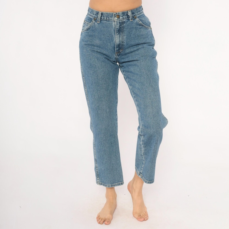 May include: Light blue denim jeans with a classic five-pocket design. The jeans have a straight leg cut and a button and zipper closure. The jeans are worn at the ankles.