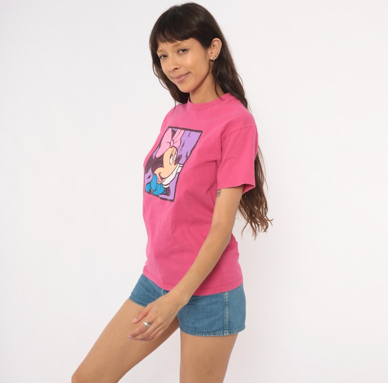 May include: A bright pink short-sleeved t-shirt featuring a rectangular graphic print of Minnie Mouse with a pink bow and blue clothing. The character is set against a purple background within a black border on the front of the cotton shirt.