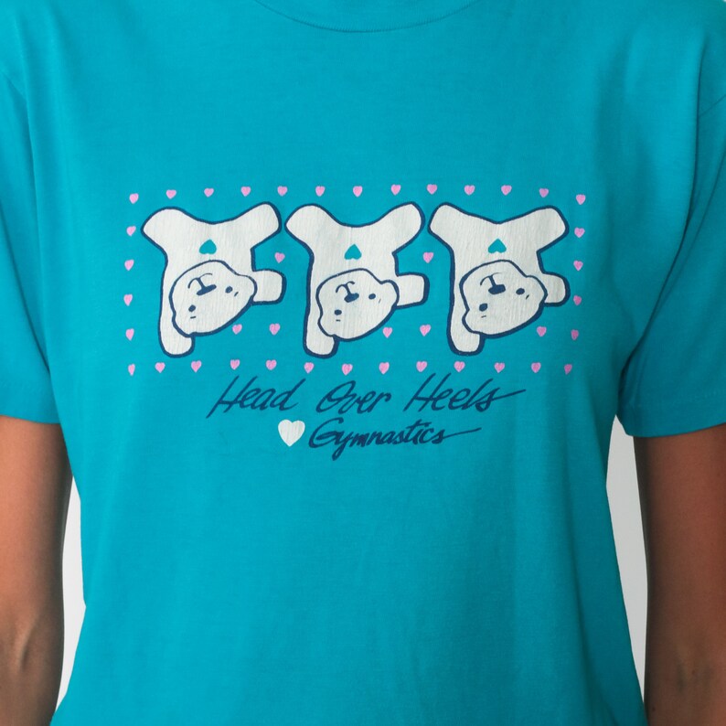 May include: A turquoise blue t-shirt with a graphic of three white bears doing a handstand. The bears are surrounded by pink hearts. The text "Head Over Heels Gymnastics" is printed below the bears.