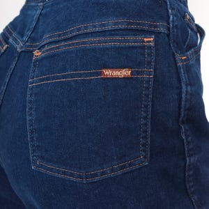 K&ouml;nnte beinhalten: Nahaufnahme einer dunkelblauen Jeans mit einer Ges&auml;&szlig;tasche. Die Tasche hat orangefarbene N&auml;hte und ein kleines rechteckiges Etikett mit dem Markennamen "Wrangler". Die Jeans ist ein klassischer Stil.