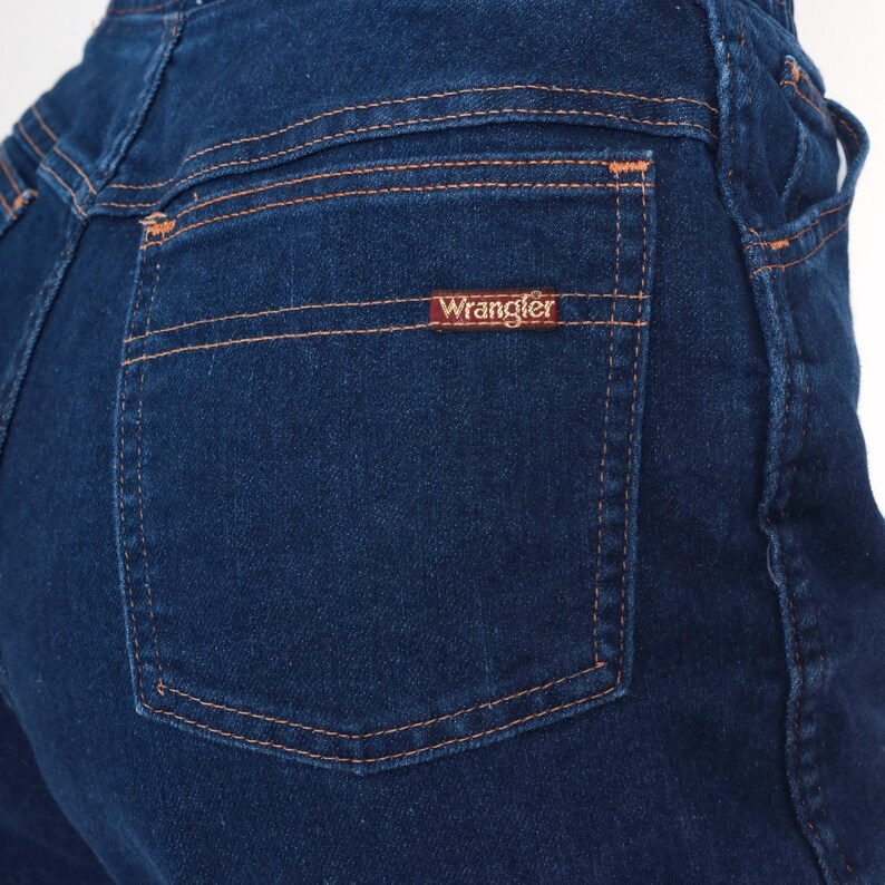 K&ouml;nnte beinhalten: Nahaufnahme einer dunkelblauen Jeans mit einer Ges&auml;&szlig;tasche. Die Tasche hat orangefarbene N&auml;hte und ein kleines rechteckiges Etikett mit dem Markennamen "Wrangler". Die Jeans ist ein klassischer Stil.