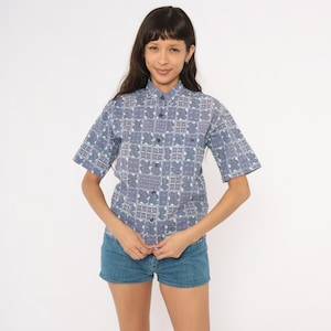 May include: A short-sleeved button-down shirt featuring a repeating grid pattern in shades of blue, white, and muted purple. The shirt has a classic collar, a front button placket, and a structured fit, paired with blue denim shorts.