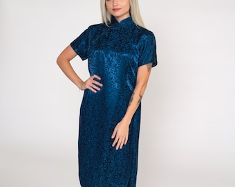May include: A teal-colored, knee-length dress with short sleeves and a mandarin collar. The dress features an all-over embossed floral pattern. The fabric has a slight sheen. The dress has a side slit and is worn by a person.