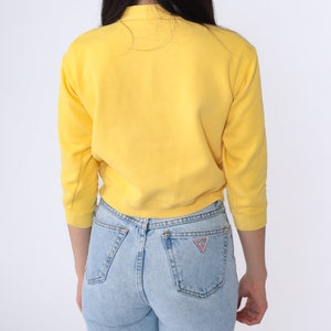 May include: A yellow cropped sweatshirt with long sleeves and a ribbed hem. The sweatshirt is worn over light blue jeans with a small triangle logo on the back pocket.