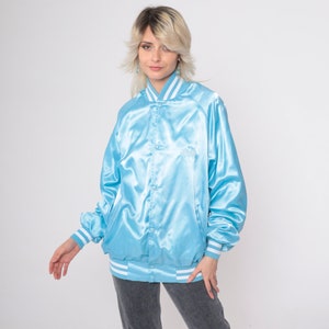 May include: A light blue satin bomber jacket with white striped trim around the collar, cuffs, and hem. The jacket has a button closure and a small embroidered logo on the chest.