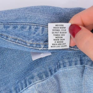 K&ouml;nnte beinhalten: Nahaufnahme eines blauen Jeanshemdes mit einem wei&szlig;en Pflegeetikett. Das Etikett enth&auml;lt Waschanweisungen: Maschinenw&auml;sche warm, Normalzyklus, nicht bleichen, Trockner mittel, warm b&uuml;geln bei Bedarf, waschen und trocknen mit &auml;hnlichen Farben.