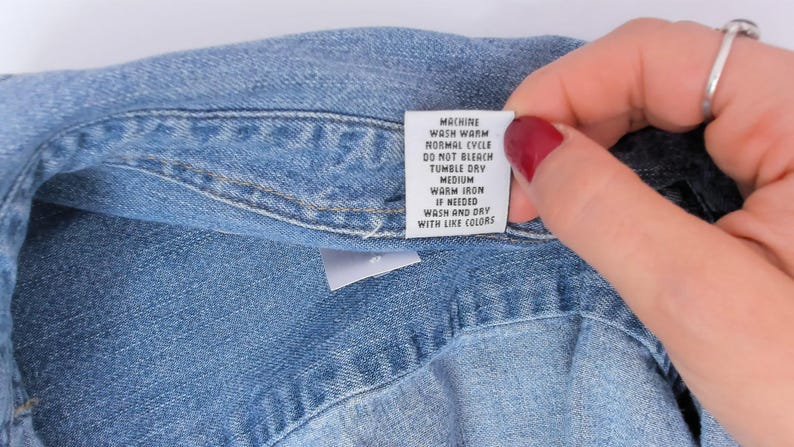 K&ouml;nnte beinhalten: Nahaufnahme eines blauen Jeanshemdes mit einem wei&szlig;en Pflegeetikett. Das Etikett enth&auml;lt Waschanweisungen: Maschinenw&auml;sche warm, Normalzyklus, nicht bleichen, Trockner mittel, warm b&uuml;geln bei Bedarf, waschen und trocknen mit &auml;hnlichen Farben.