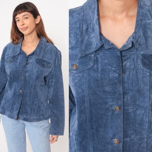 May include: A blue suede jacket with a button-up front, two chest pockets, and a collared neckline. The jacket has long sleeves and is a medium shade of blue. The buttons are a bronze color.