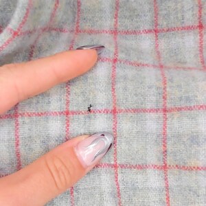 Puede incluir: Una tela gris claro con un patr&oacute;n de cuadr&iacute;cula roja. El material tiene una apariencia suave y texturizada con una peque&ntilde;a imperfecci&oacute;n oscura y singular visible en la superficie del textil, resaltando el tejido y las l&iacute;neas rojas.