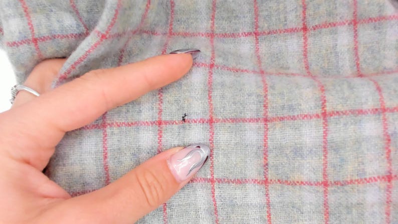 Puede incluir: Una tela gris claro con un patr&oacute;n de cuadr&iacute;cula roja. El material tiene una apariencia suave y texturizada con una peque&ntilde;a imperfecci&oacute;n oscura y singular visible en la superficie del textil, resaltando el tejido y las l&iacute;neas rojas.