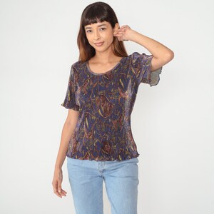Puede incluir: Una camisa de manga corta azul y dorada con estampado y cuello redondo.