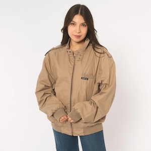 May include: A tan Members Only jacket featuring a full-length front zipper, a high collar with snap buttons, shoulder epaulets, and ribbed cuffs. The jacket includes a small chest pocket with a black label displaying white text.