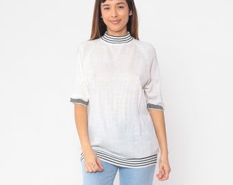 May include: White mock neck sweater with black striped trim on the collar, sleeves, and hem. The sweater is made of a soft, lightweight fabric.