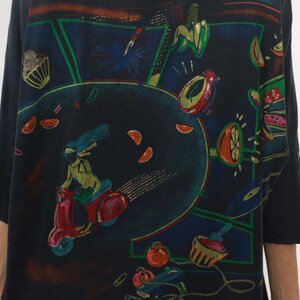 May include: Black long-sleeve shirt with a colorful, retro-style graphic. The design features a person riding a red scooter, surrounded by fruit, a cupcake, and other whimsical elements. The artwork has a vibrant, vintage aesthetic.