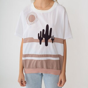 May include: White short-sleeved shirt with a brown and white striped pattern and a black cactus graphic. The shirt has a round neckline and a relaxed fit.