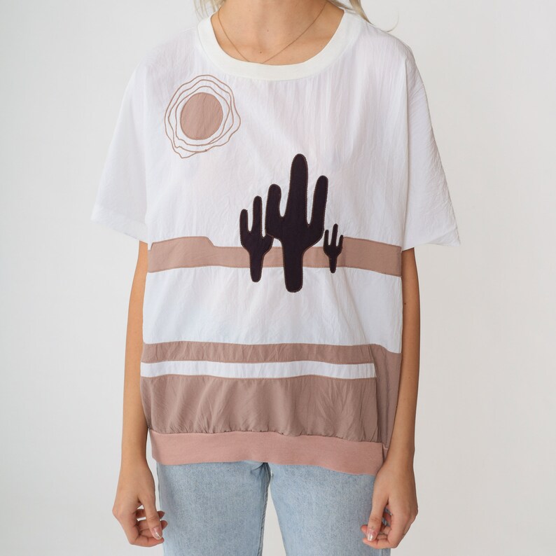 May include: White short-sleeved shirt with a brown and white striped pattern and a black cactus graphic. The shirt has a round neckline and a relaxed fit.