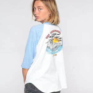 May include: White and light blue raglan-sleeve t-shirt featuring a graphic of the USS Independence aircraft carrier on the back. The graphic includes the ship, ocean, sun, and text. The shirt has a relaxed fit.