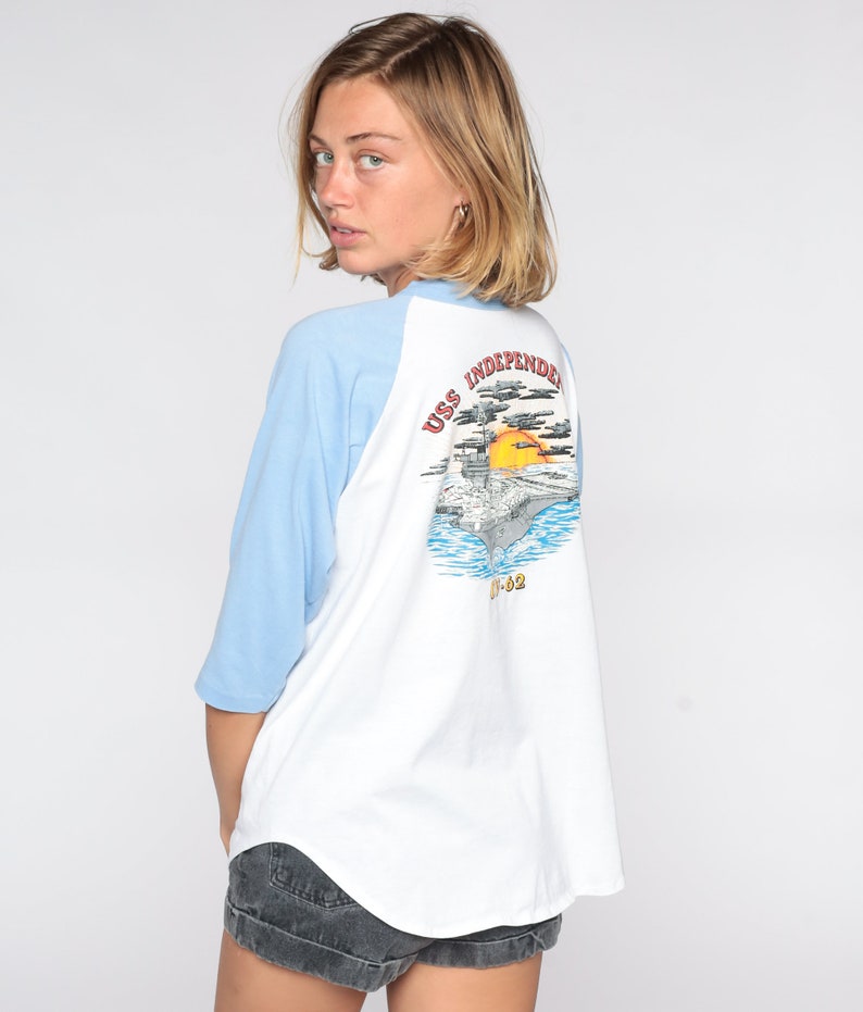 May include: White and light blue raglan-sleeve t-shirt featuring a graphic of the USS Independence aircraft carrier on the back. The graphic includes the ship, ocean, sun, and text. The shirt has a relaxed fit.
