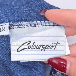 Puede incluir: Una prenda de mezclilla azul con una etiqueta de tela blanca que presenta l&iacute;neas horizontales finas y el nombre de la marca "Coloursport" impreso en negro. Una etiqueta blanca m&aacute;s peque&ntilde;a en el lateral muestra el n&uacute;mero 12 en negro.