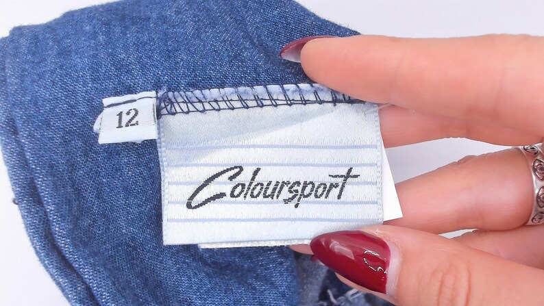 Puede incluir: Una prenda de mezclilla azul con una etiqueta de tela blanca que presenta l&iacute;neas horizontales finas y el nombre de la marca "Coloursport" impreso en negro. Una etiqueta blanca m&aacute;s peque&ntilde;a en el lateral muestra el n&uacute;mero 12 en negro.