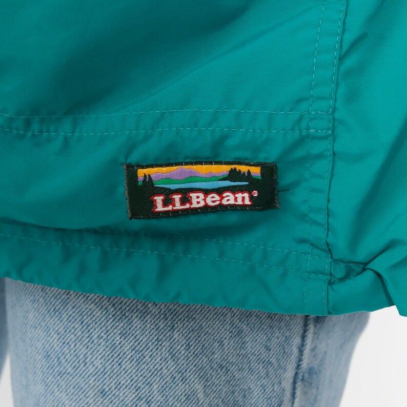 May include: A teal blue jacket with a sewn-on patch that reads "LLBean" in red letters with a mountain scene behind the text.