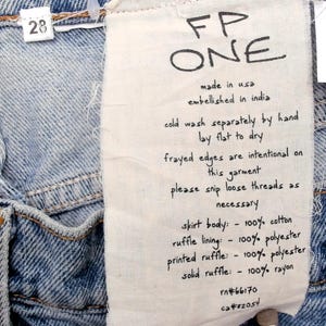May include: Close-up of light blue denim jeans with a white tag. The tag reads "FP ONE" and includes care instructions and fabric composition details. The jeans have a size 28 tag.