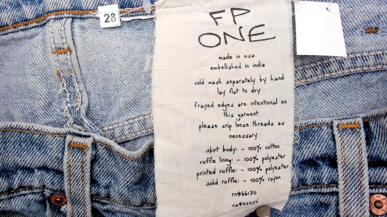 May include: Close-up of light blue denim jeans with a white tag. The tag reads "FP ONE" and includes care instructions and fabric composition details. The jeans have a size 28 tag.