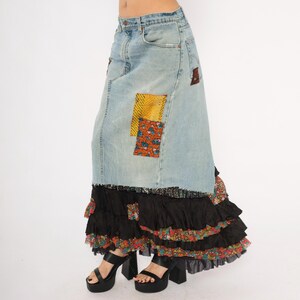 May include: A light-wash denim skirt with patchwork details. The skirt has a high waist and a long, flowing silhouette. The bottom of the skirt has layers of black ruffles with floral accents. The model is wearing black platform heels.