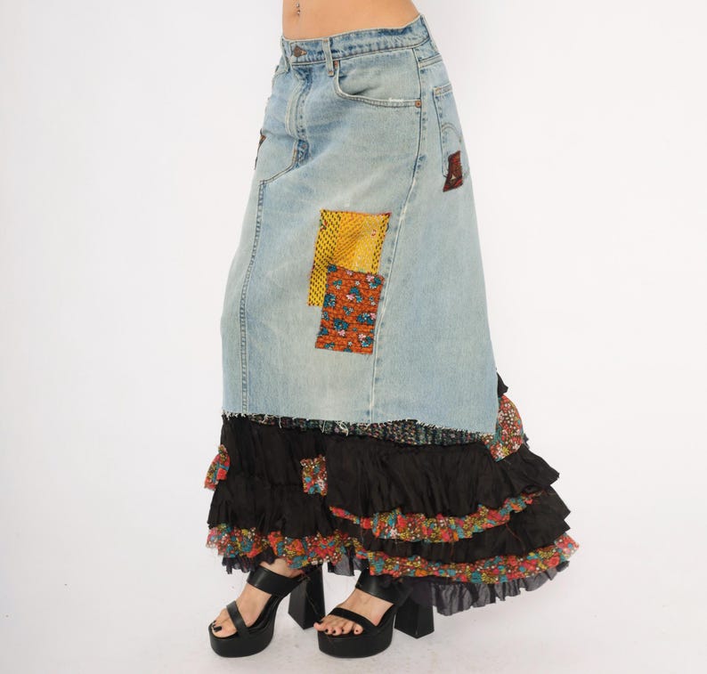 May include: A light-wash denim skirt with patchwork details. The skirt has a high waist and a long, flowing silhouette. The bottom of the skirt has layers of black ruffles with floral accents. The model is wearing black platform heels.