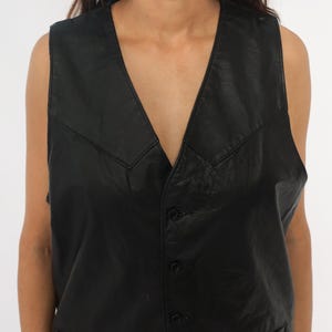 May include: A black leather vest with a V-neckline and button closure. The vest has a classic design with a tailored fit. The leather appears to be slightly textured, adding visual interest. The vest is sleeveless and suitable for layering.