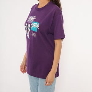 May include: A purple t-shirt with a crew neck and short sleeves. The shirt features a graphic design with the words "CAMP" and "CEMBRIDGE" in white and teal. The shirt is worn with light blue jeans.