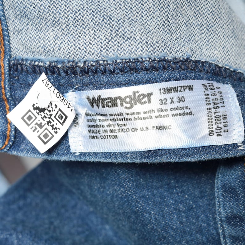 May include: A blue denim Wrangler jeans label with white text. The label says "13MWZPW 32 X 30" and includes care instructions. The text says "Machine wash warm with like colors, only non-chlorine bleach when needed, tumble dry low. Made in Mexico of U.S. Fabric 100% Cotton."
