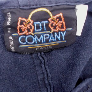 May include: Close-up of a navy blue garment tag. The tag features the text "DT COMPANY PARIS" in blue and gold embroidery. The tag also has the text "FABRIQUE EN FRANCE" in white. The tag has two orange floral designs.