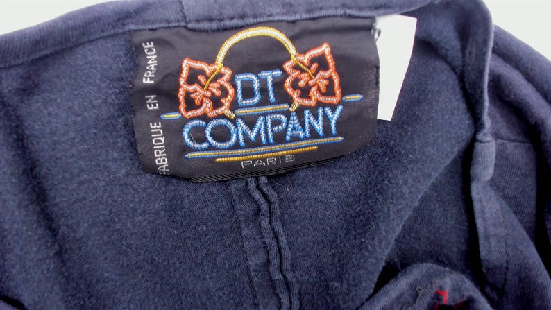 May include: Close-up of a navy blue garment tag. The tag features the text "DT COMPANY PARIS" in blue and gold embroidery. The tag also has the text "FABRIQUE EN FRANCE" in white. The tag has two orange floral designs.