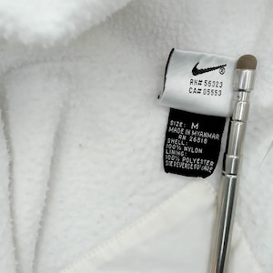Puede incluir: Primer plano de una chaqueta de forro polar blanca con una etiqueta Nike. La etiqueta incluye el logotipo de Nike, los n&uacute;meros RN y CA, la talla M e informaci&oacute;n sobre el material. Tambi&eacute;n se ve un l&aacute;piz t&aacute;ctil plateado.