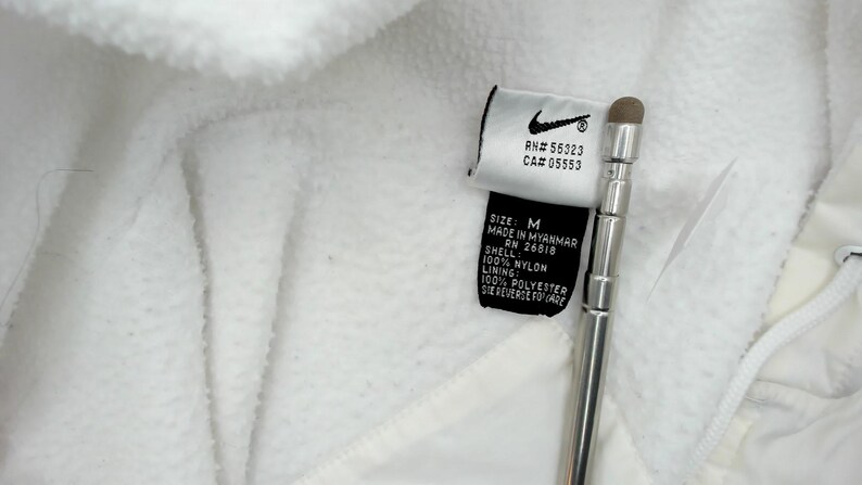 Puede incluir: Primer plano de una chaqueta de forro polar blanca con una etiqueta Nike. La etiqueta incluye el logotipo de Nike, los n&uacute;meros RN y CA, la talla M e informaci&oacute;n sobre el material. Tambi&eacute;n se ve un l&aacute;piz t&aacute;ctil plateado.
