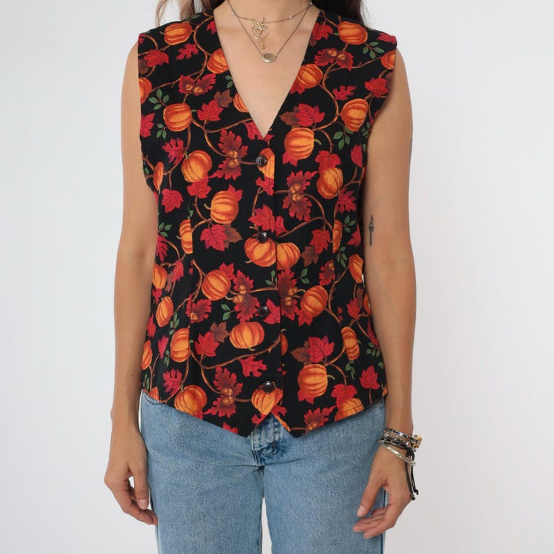 Puede incluir: Un chaleco negro sin mangas con estampado de calabazas y hojas. El chaleco tiene cuello en V y cierre de botones.