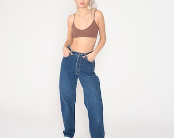 May include: A woman wearing a brown ribbed tank top and blue denim jeans with a high waist. She is standing with her hands in her pockets.