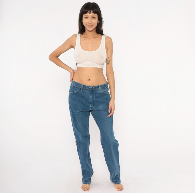 May include: A light-colored crop top and blue jeans. The crop top is a neutral color, and the jeans are a medium wash denim. The jeans have a classic five-pocket design and a straight leg fit. The model is standing in front of a white background.