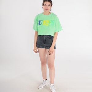 May include: A green cropped t-shirt with a faded "GUESS" logo in yellow, blue, and green. The model is wearing the shirt with black denim shorts and white trainers.