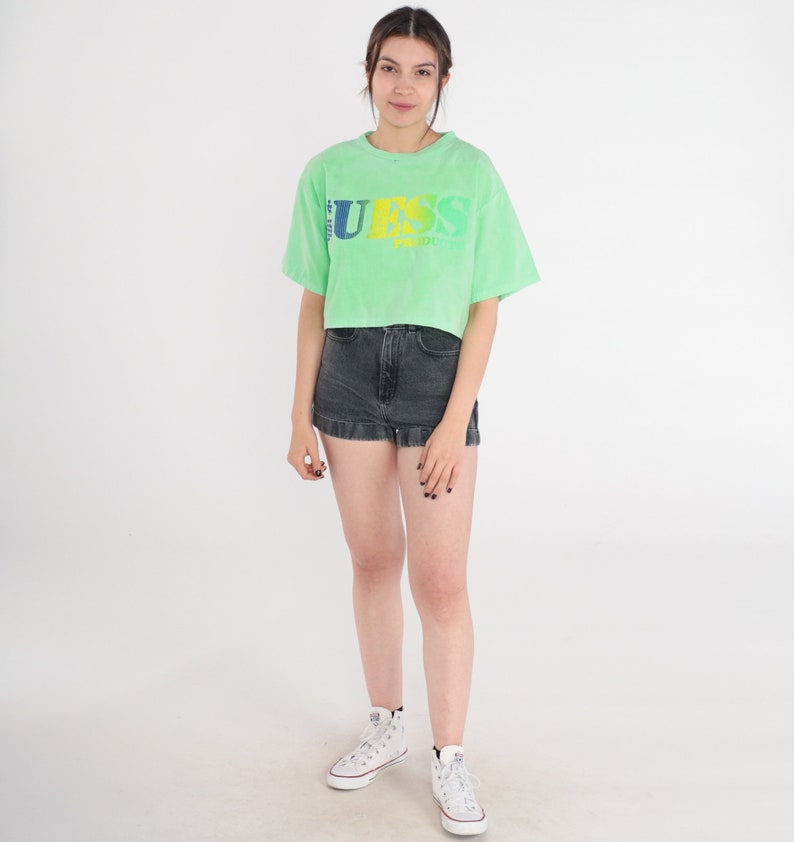 May include: A green cropped t-shirt with a faded "GUESS" logo in yellow, blue, and green. The model is wearing the shirt with black denim shorts and white trainers.